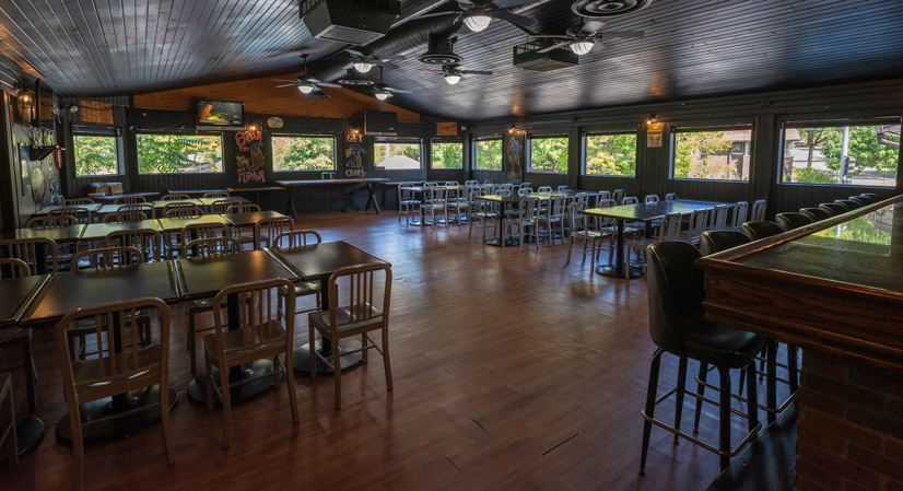 A large space in a restaurant, wood flooring and fans on the ceiling, there are tables and chairs lined up, and large windows along the walls revealing trees around the building.
