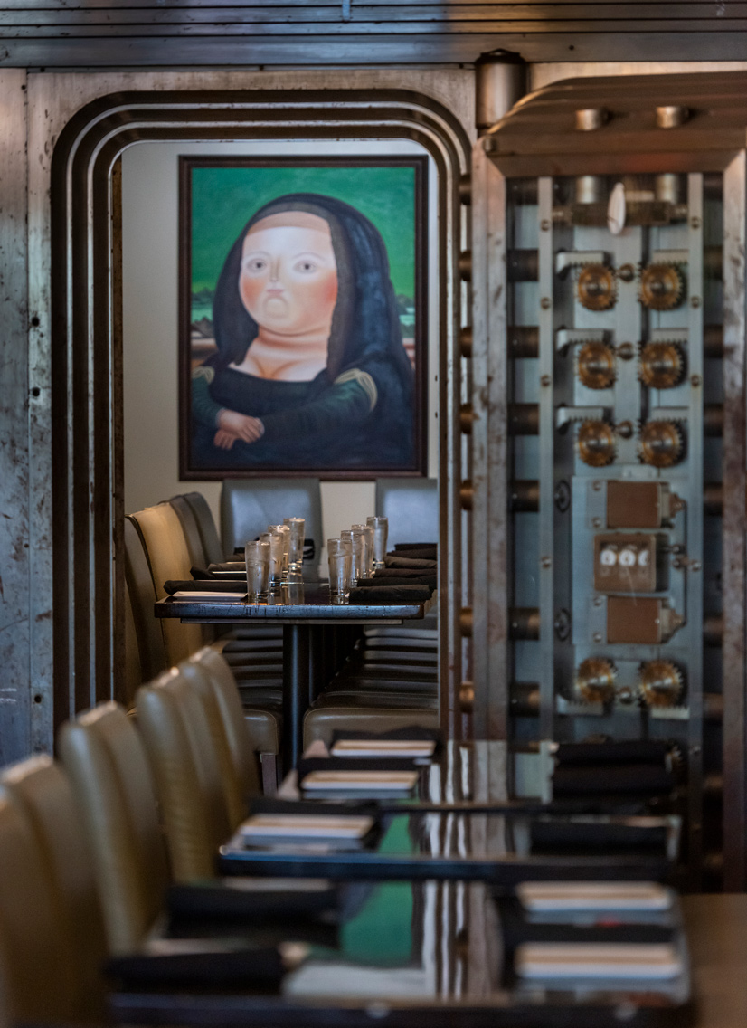 tables with reflective surface and chairs lined up. The inside of a vault door reveals gears for locking the door, inside the door is more tables and chairs lined up and a recreated painting of the Mona Lisa on the wall.