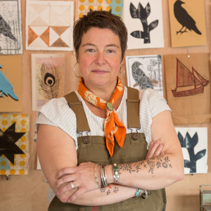 woman with short hair and overalls and arms crossed standing in front of quilt