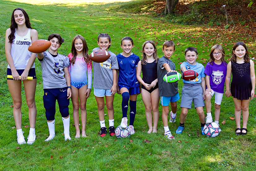a teen and 9 children lined up in various sport uniforms in the fall outdoors