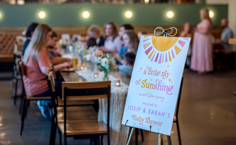 people dining at tables in a restaurant, in the foreground there is a sign that reads "a little ray of sunshine is on the way, welcome to Josip & Sarah's Baby Shower"