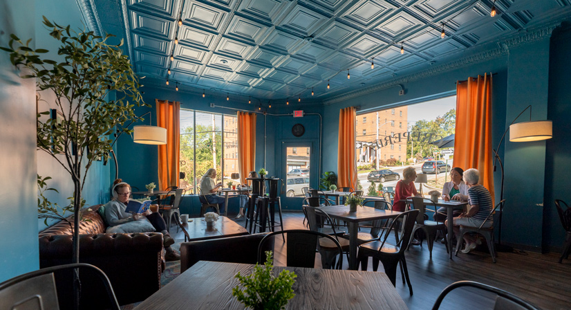 A medium sized room with a counch and some tables and chairs sprawled out. There are two large windows with orange curtains, and the walls are a turquise blue