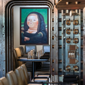 tables with reflective surface and chairs lined up. The inside of a vault door reveals gears for locking the door, inside the door is more tables and chairs lined up and a recreated painting of the Mona Lisa on the wall.