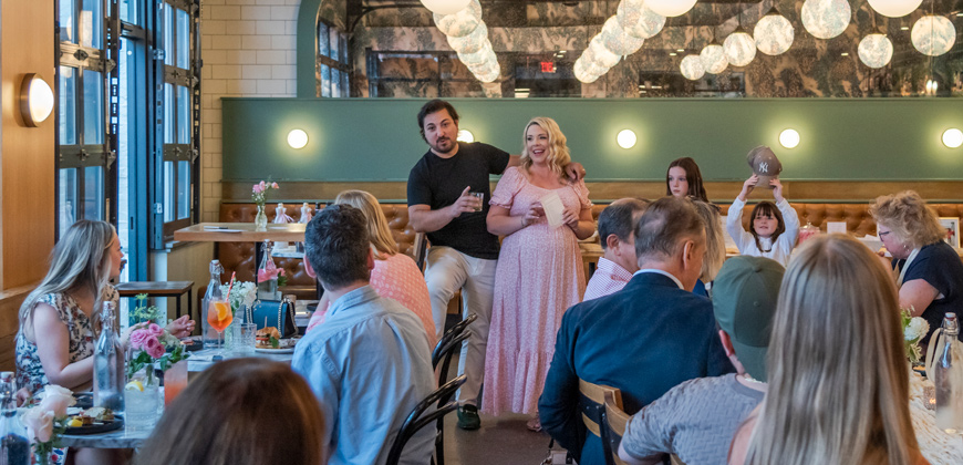 two people, a a man and a woman, standing among people sitting at tables with food, giving a speech