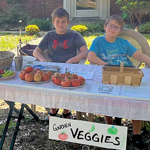 two boys sit at a table with produce. The boy on the right has glasses.