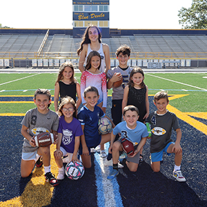 10 kids posing on a football field holding footballs and soccer balls