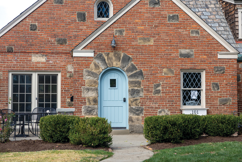 A red brick house, with large grey stones sparsely placed throughout. A walkway leads up to the front door, with shrubs lined up on each side. On the right side of the house is a window, with a white window pane underneath with the number 101 on it. The door is a light sky blue door, and the trim around it has large stone bricks around it. The top of the door is curved with a small window near the top. The door handle is black.