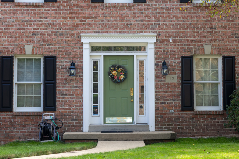 A red brick house with a concrete walkway leading up to a small porch in front of the door. There are two windows on the side with black shutters. The doorway trim is a decorative white, with small windows around each side of the door and the top of the door. The door is an olive green color with a gold handle, in the middle of the door is a fall wreath. On both sides are two sconce porch lights, on the right side is the house number 112.