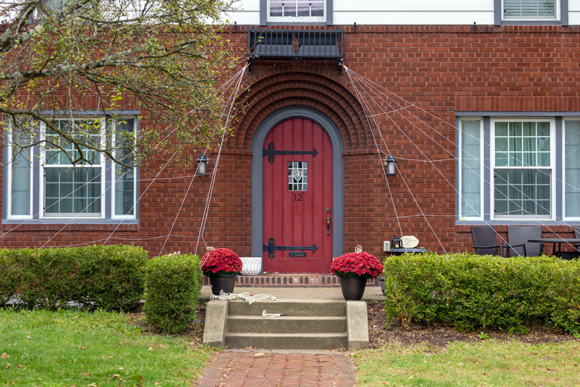 the entrance of a red brick house, a red wooden door is inset from the brick, with a blueish grey trim. The door has larbe black hinges that extend past the middle of the door. In the center of the door is a small glass window with the number 12 below it. there is a small concrete porch with steps leading to the door, two flower pots with red flowers sit on the porch next to rows of bushes extending to the side of the house.