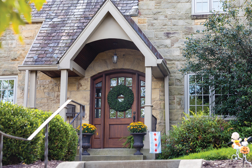 A grey large stone brick house with steps leading to the front door. On the left side is an iron railing and some bushes. Above the door is a light tan colored awning, and a porch light is hanging from it underneath. On each side of the door is potten plants with yellow flowers. The door is a walnut wood color with 4 windows on each side of the door trim. The door has several small glass panels partially covered by a large green wreath.