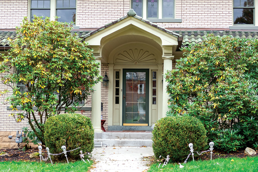 A light tan colored brick house, there is a concrete pathway leading to the front porch of the house, on each side are small shrubs, and some large greenery obstructing the view of the windows. There are two steps up to the porch, with two large pillars on each side of the walkway, holding up a small roof with slate tiles.The door has light tan decorative trim, on each side are for small windows, and above the door is a decorative half circle, sun burst decoration. The door is dark green, and there is a dark green wreath on it.