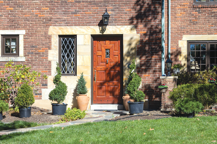 A red brick house, with a stone path leading up to the front door. There are small potted bushes surrounding the front door. A small brick pillar on the right has a black lamp post on it. The front door has Large light colored bricks around it, and a window to the left of the door. Above the door is a sconce style porch light. The door is a reddish color wood with several panels. In the middle is a stained glass window, and the handle is a decorative metal piece.