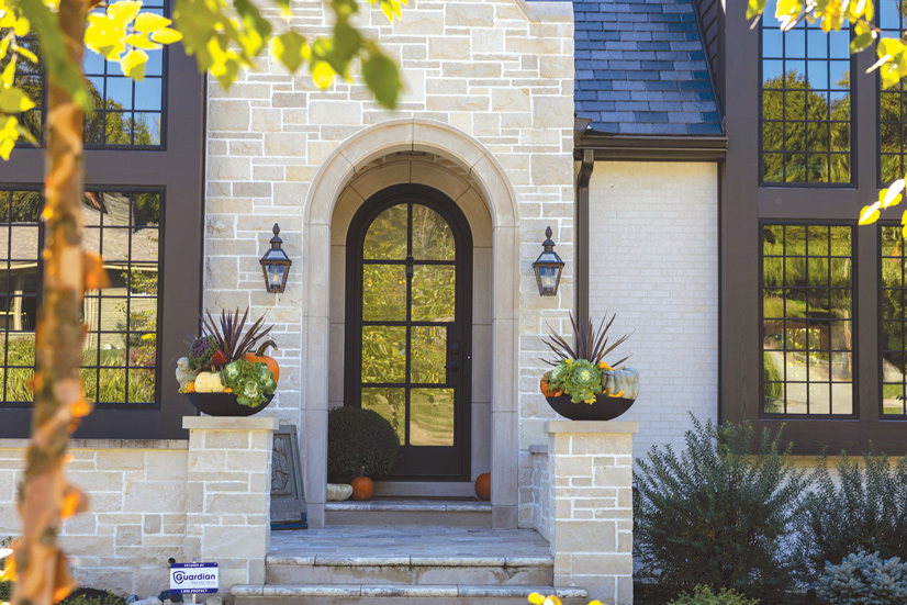 a light colored brick house, with large bricks. There are steps leading to the front door, there are two short pillars with decorative bowls on each side, there are flowers and pumpkins in them. On each side of the door is two sconce lamps. The front door is inset from the front of the house, inside is two small pumpkins and a large flower pot. The door is a dark wooden frame, rounded at the top. The door is mostly glass windows that is refecting the greenery from the yard.