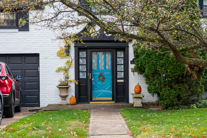 A brick house painted white, there is a tree in the front yard with overhanging branches. A concrete walkway leads to a small porch. There is a stone bench with a pumpkin on it, and a large stone flower pot with flowers coming out, and a pumpkin next to it on the porch. The front door is lined with black decorative trim, and several windows that have a frosted decoration on it that looks like large leaves. The door is a bright cyan farmhouse door, with three vertical windows on the top showing the same frosted decoration. In the middle of the door is a red wreath. To the right of the door is a large bush covering up the house number and mail box.