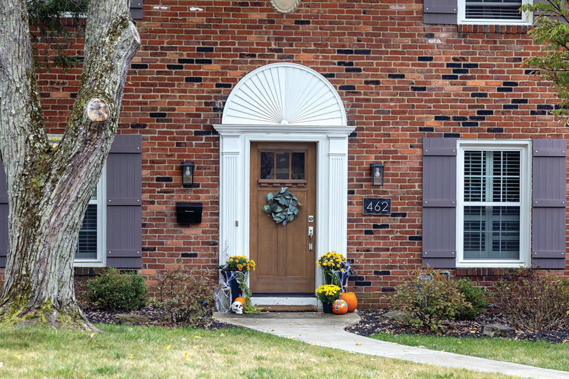 A red brick house with black bricks sparsely placed. The windows have white trim and priple shutters. A concrete walkway leads to the front door, on each side is bushes starting to lose their leaves. In front of the door is a display of yellow mums and pumpkins. The door has a decorative white trim, above the door is a half circle with decorative lines leading to the center. The door is a medium wood color with a silver door handle. At the top of the door is glass pane, and underneath that is a dark green wreath with large leaves.