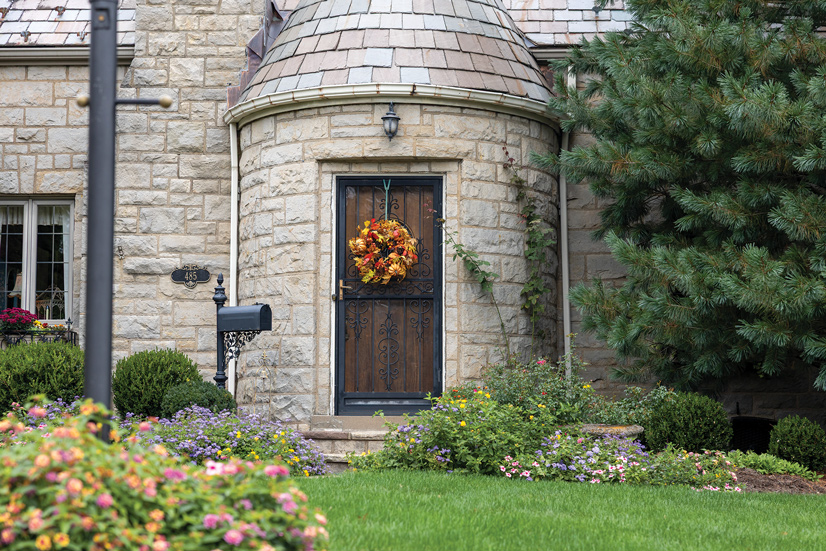 a large stone house with many flowers lining the outside. A large pine tree is on the right side of the house. The entrance way is a rounded pillar, and there is a black metal mailbox to the left of the door, next to that is the house number 485. There is an black metal screen door with decorative flourishes throughout, and hanging in the middle is a yellow and orange fall wreath. Behind the screen door is a dark wood door.