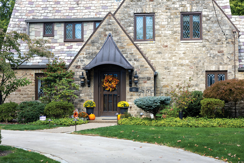 A large brick stone cottage style house, with an entry way that is extruding from the front of the house. There is a curved driveway and a small pathway leading to the front of the house, the front of the house is lined with greenery and many different sized bushes and shrubs. On both sides of teh door is a potted plants with yellow flowers and pumpkins. The door itself has a dark wood frame, and above is a dark colored, triangular awning that slopes down. On each side of the awning are sconce style porch lights, and on the right side is the number 83 on a black metal plate. The dark wood door has a brightly colored orange and red wreath on it.