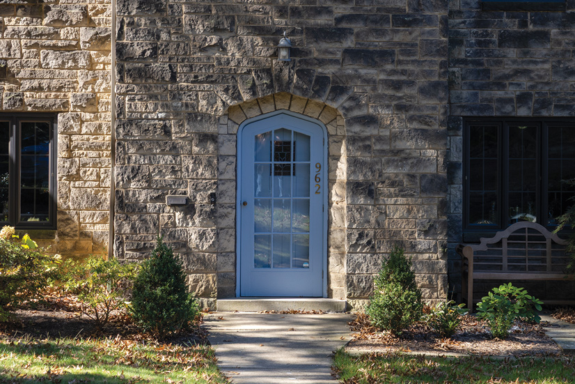 a rough textured stone house, there are small shrubs in front and a decorative wooden bench in front of one of the windows. A concrete pathway leads to the front door. The door is inset into the stone and has a decorative rounded top that comes to a centrical point. There is a white screen door with a large window, on the right side of the door trim is the house number 962 in gold. Behind the screen door is a white wooden door with a decorative glass window