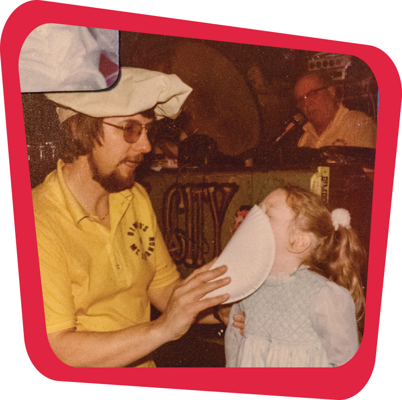 An old photo of a man in a hat, glasses and a yellow shirt, throwing a pie in the face of a little girl