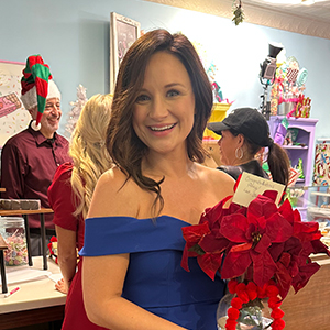 A woman with dark brown hair wearing an formal off the shoulder royal blue dress holds a bouquet of red poinsettias. Three people dressed in red and green can seen talking in the background.