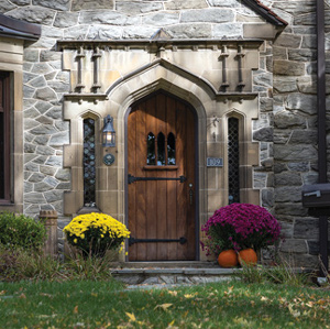 A grey stone house with a castle like appearance. There is a step leading up to the door entryway, on each side of the door is potted flowers, one with yellow and one with magenta flowers, There are two decorative pumpkins as well. The trim around the door is a greyish tan stone decoration. There are two long glass stained windows on each side, on the left is a lamp post, on the right is the number 109 on an iron plate. The door itself is wood, with two large black metal hinges reaching across the front. There are three small windows in the center that have a point at the top.
