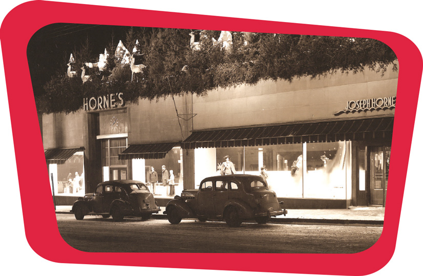A black and white photo of Horne's department store, its a concrete building, with pine trees on top. Below the awnings are large glass windows with mannequins sporting vintage clothes. Two cars sit on the street indicating the time is in the 1940s
