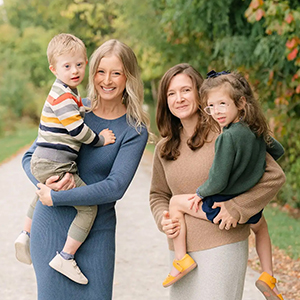 Two women standing together outdoors, each holding a child.