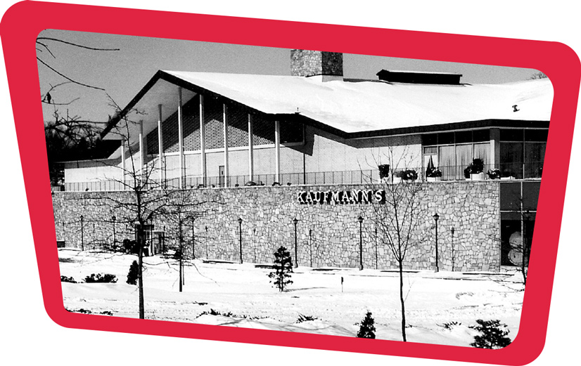 A black and white photo of Kaufmans department store, there is a large cobblestone wall, with "Kaufmann's" in lettering on it. The parking lot is empty and snow covered. The building peaks out from behind the wall with a large pointed roof and pillars lining the front. Above is a large chimey sticking out of the middle of the building.