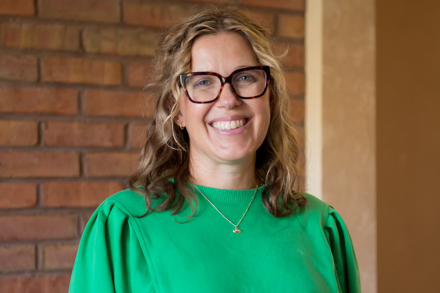 A woman with light brown shoulder length hair and dark framed glasses wearing a bright green long sleeve top stands smiling in front of a red brick wall.