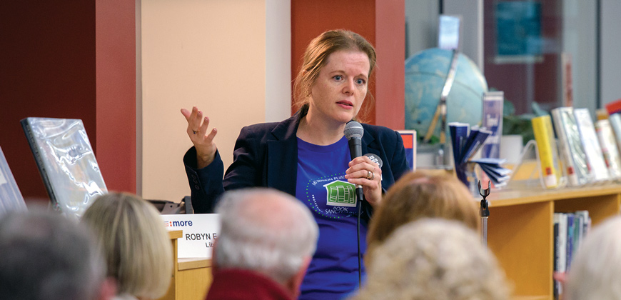 A blonde woman wearing a blue shirt and black blazer holding a microphone speaking to a group of people