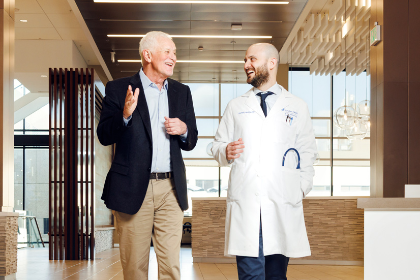 two men walking through the lobby of a hospital, one man has a beard and is wearing a white doctors coat. The other is older and wearing a black blazer and khakis.