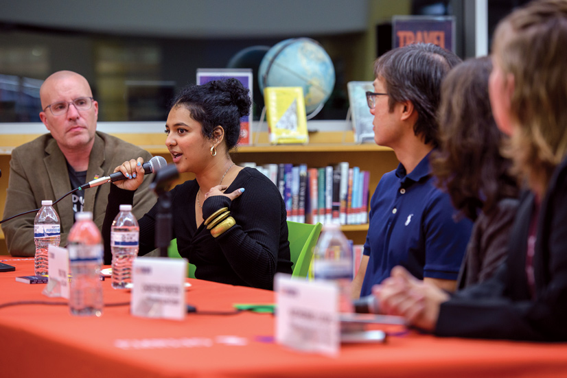 A dark haired woman holding a microphone sitting at a table of people with name tags speaking to a group of people