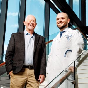 two men walking through the lobby of a hospital, one man has a beard and is wearing a white doctors coat. The other is older and wearing a black blazer and khakis.