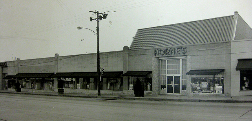 A black and white photo of HORNE'S department store. Its a large concrete building along the street. Large windows are surrounding the entrance to the building with a large neon sign that sasys "HORNE'S" above it. The rest of the storefront is lined with display windows with mannequins and other items on display.