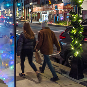 Two people walking away from the camera together on a sidewalk dressed in winter coats and hat with one carrying a brown paper shopping bag. Holiday decorations and building exteriors can be seen in the background.