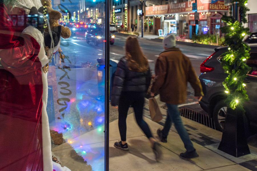Two people walking away from the camera together on a sidewalk dressed in winter coats and hat with one carrying a brown paper shopping bag. Holiday decorations and building exteriors can be seen in the background.