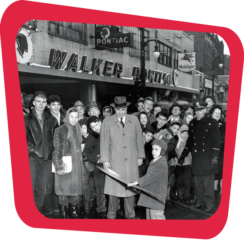 A black and white photo, there is a large number of people gathered outside a building with a neon sign above that reads "WALKER PONTIAC". A man wearing a coat and hat is supervising a child holing a large ribbon about to be cut.
