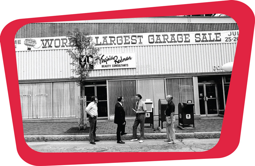 A black and white photo of the front of a building with a sign that says "Worlds Largest Garage Sale" in western typography. Below that is a sign that says "virginia redman beauty consultants". There are4 men standing on the street and sidewalk in front of the building. There are several large windows along the sidewalk, covered with curtains.