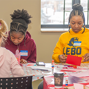 teenager wearing a burgundy hoodie and woman wearing a bright yellow sweatshirt with large navy block letters spelling out LEBO sit together at a table making cards out of red and pink paper.