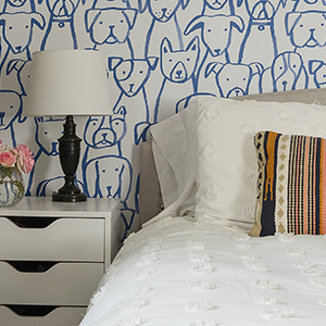 A bed with a fluffy white comforter and a striped pillow with a white nightstand to the left. Illustrated wallpaper in the background features whimsical drawings of dogs in bright blue.