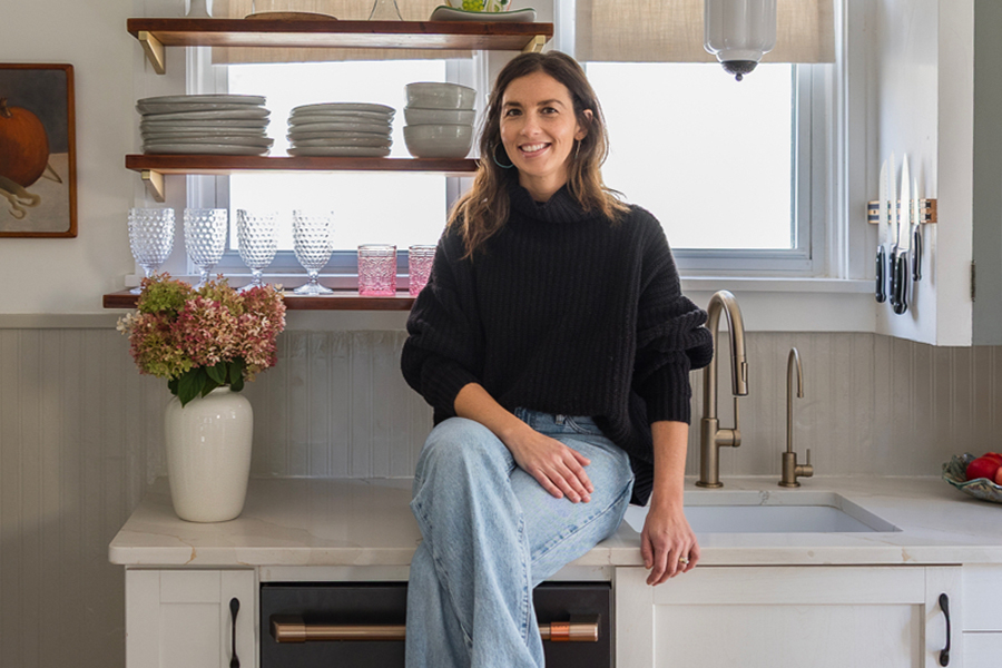 A women with long brown hair wearing an oversized black turtleneck sweater and jeans smiles for the camera while sitting on the counter in a kitchen. A large window with shelves holding a variety of cups, plates and bowls can be seen in the background.