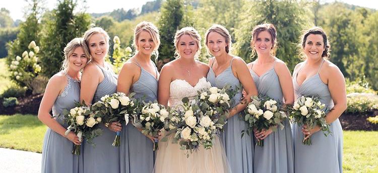 a portrait of several women, 6 bridesmaids in pale blue dresses and one bride in a white wedding dress lined up posing holding white flowers, in a field in front of trees