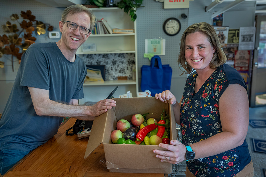 Two people standing across from each other propping up a cardboard box filled with a variety of fresh produce.