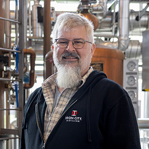 A man with short grey hair and beard wearing a navy blue sweatshirt smiles for the camera.