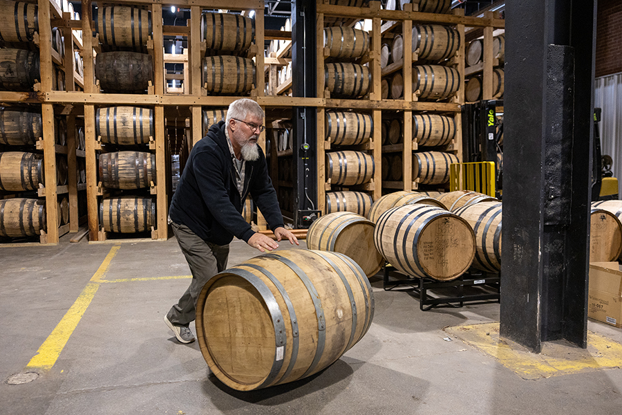 A man with short grey hair and beard wearing a navy blue sweatshirt rolls a large wooden barrel through a warehouse.