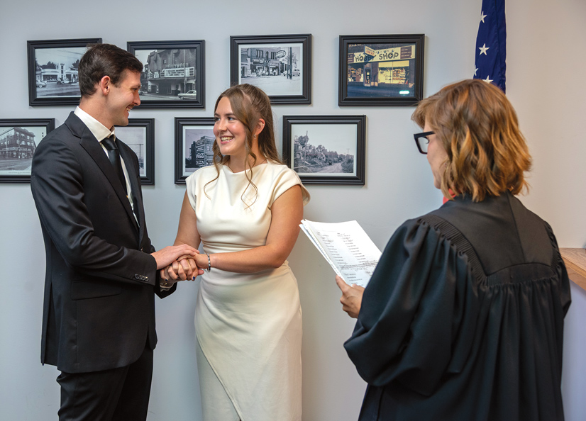 A man in a black suit and a woman in a white dress standing next to each other looking at each other holding hands, another woman wearing a judges robe is standing next to them holding paper, some historical photos are on the wall behind them.