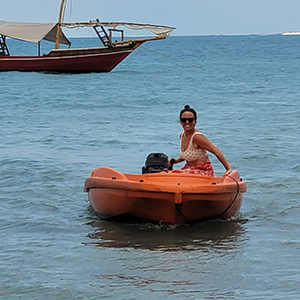 A woman sitting in a small orange motorboat navigating it towards the camera. The ocean an sailboat can be seen in the background.