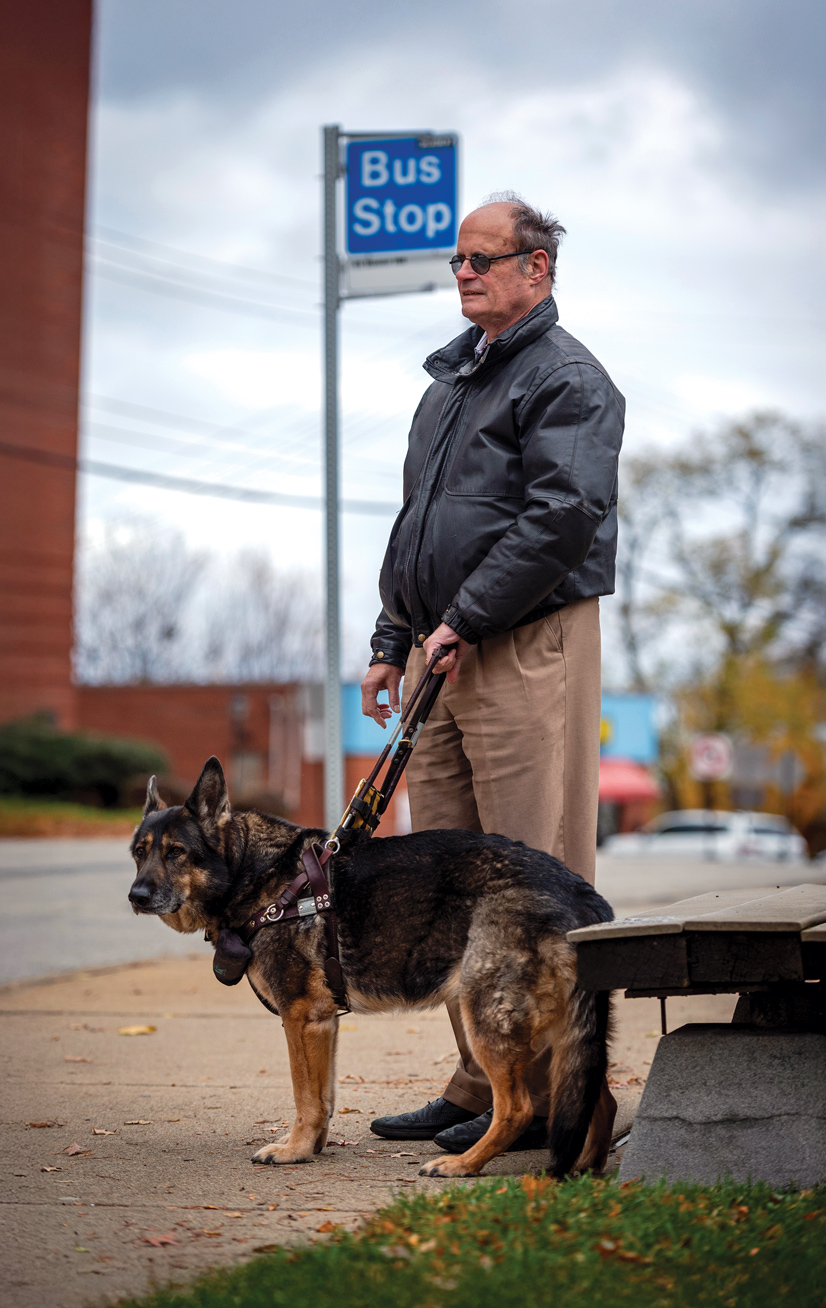 A man wearing sunglasses and a black coat waiting at a bus stop with his seeing-eye-dog
