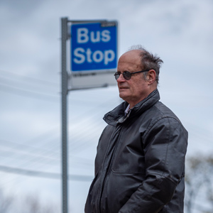 A man wearing sunglasses and a black coat waiting at a bus stop with his seeing-eye-dog