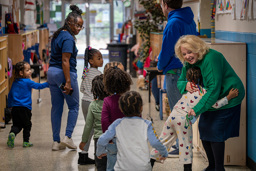 A busy school building hallway with several children dressed in colorful outfits walking in a line away from the camera. One student is hugging a woman dressed in a bright green sweater and dark skirt.